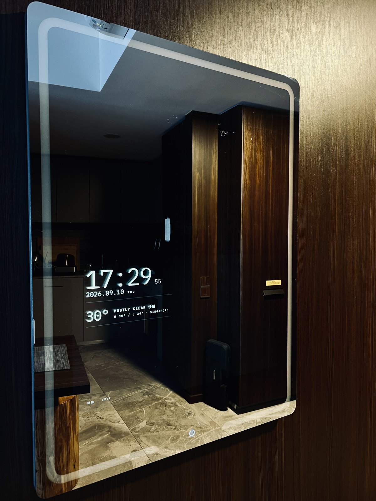 The mirror on a wood-paneled hallway wall in daylight, its frame lit, showing only the time, date, and weather over the reflection of the room