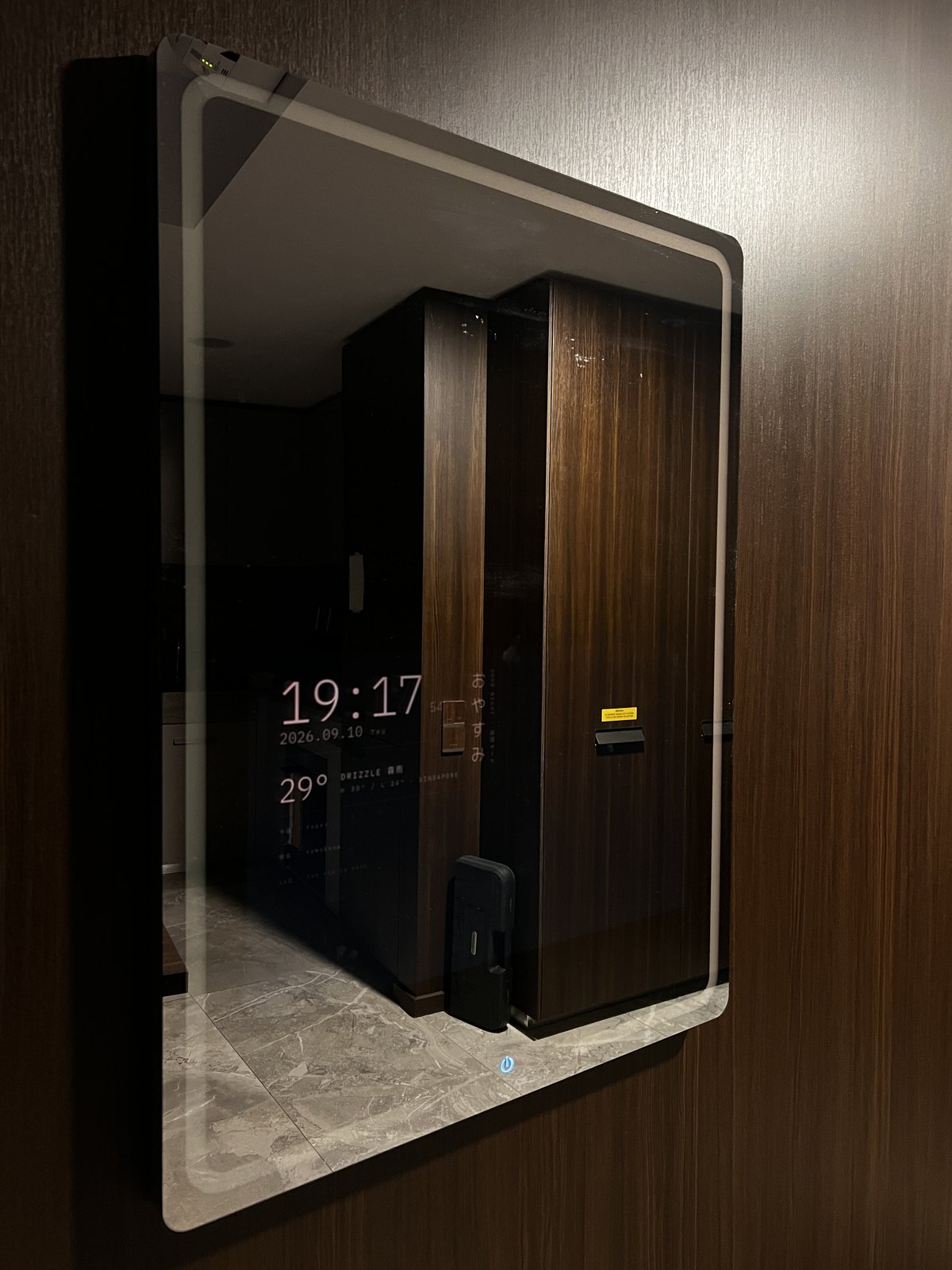 The mirror at night on the same wall: the frame's light ring on, the clock, date, and weather in dim red over the reflection of the hallway